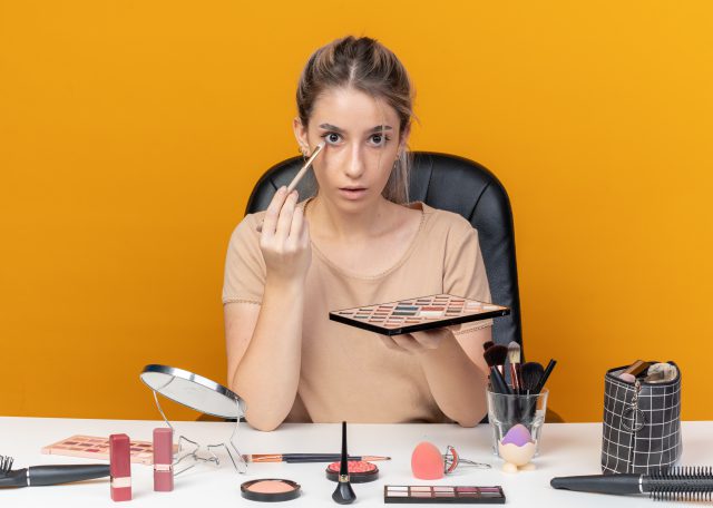 surprised young beautiful girl sits at table with makeup tools applying eyeshadow with makeup brush isolated on orange background